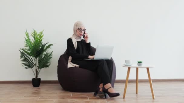 ビーンバッグでポータブルコンピュータを活用しながらビジネスコールを持っているヒジャブの陽気な女性 コピースペースで職場での商業コミュニケーションに成功する喜ばしいアラブ起業家 — ストック動画
