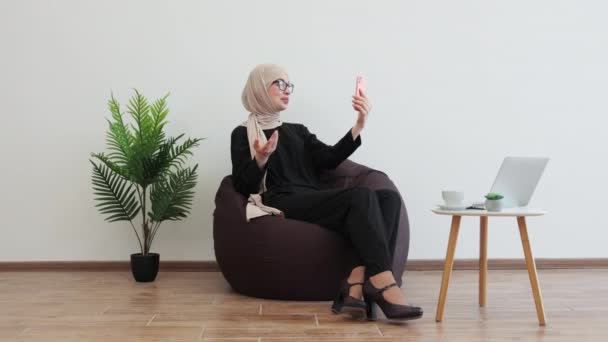 微笑的女性穿着穆斯林的正式服装在手机上记录视频而坐在书包椅子上 复制空间 愉快的商务女士在头巾分享成功谈判的经验 — 图库视频影像