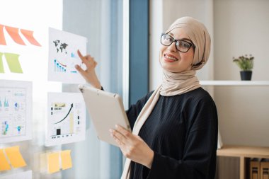 Dijital tabletli çekici Arap bayan iş yerindeki şema ve çizelgeleri kullanarak finansal istatistikleri inceliyor. Akıllı aygıttaki bilgileri kağıt raporuyla eşleştiren yetkili veri analisti.