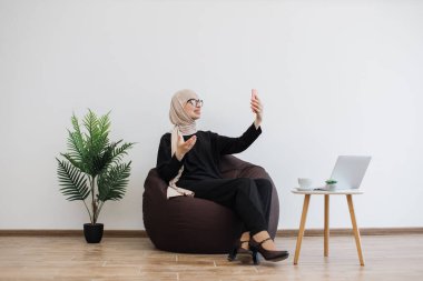 Muslimah resmi kıyafet içinde gülümseyen kadın cep telefonuyla video kaydederken pouf sandalyesinde oturuyor, fotokopi çekiyor. Başörtüsü paylaşma deneyimi olan başarılı iş kadını..