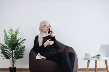 Kendine güvenen, tesettürlü, kendine güvenen, önemli telefon konuşmaları yapan ve fotokopi alanının yanında rahat bir şekilde oturan Arap bir kadın. Gözlüklü dikkatli işletme sahibi yoğun program sırasında anlaşmaları tartışıyor.