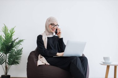 Muslimah resmi kıyafetli hoş bir genç kadın akıllı telefondan konuşuyor ve puf minder dizüstü bilgisayar kullanıyor. Kopya alanı ile iş yerinde müşteriyle önemli bilgi alışverişi yapan tesetteki iş adamı.
