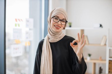 Gözlüklü gülümseyen Arap yetişkin portresi ahşap masanın kenarına oturmuş, elleri göğsünde katlanmış, işaret veriyor.