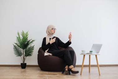 Muslimah kıyafetli neşeli genç bayan cep telefonunu fotokopi alanında tutarken selam veriyor. Çalışma saatleri boyunca el bilgisayarı üzerinde video çağrısı almak memnuniyet verici..