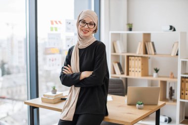 Elleri göğsünde katlanmış, ahşap masanın kenarında oturan, göz giysisi giymiş gülümseyen Arap yetişkin portresi. Bej tesettürlü neşeli iş kadını modern işyerinden başarılı bir şirket yönetiyor..