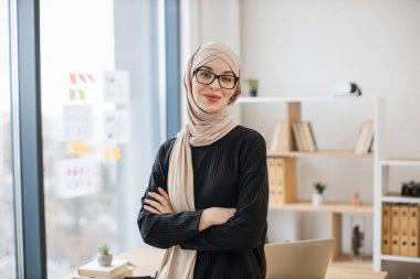 Yaratıcı iş yerinde kollarını kavuşturmuş geleneksel türbanlı çekici Müslüman bayanın yakın görüntüsü. Gözlüklü etkin girişimci parlak ofiste profesyonel faaliyetler yürütüyor..