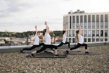 Spor kıyafetleri içinde, Asana 'da, çakıllı arazide lastik paspas kullanarak çalışan sağlıklı genç kadınlar. Çekici beyaz kadınlar güzel şehir manzarasının tadını çıkarırken denge ve duruşu geliştiriyorlar..