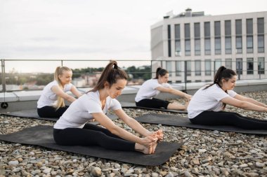 Beyaz tişörtlü ve siyah pantolonlu bir grup kadın çatıda kalırken öne doğru eğiliyor. Genç yoga pratisyenleri dışarıda antrenman yaparken beden ve zihin dinginliği hissediyorlar..