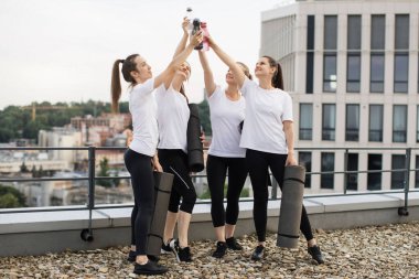 Lastik hasırlı çekici kadınların çakıllı çatıda dururken spor şişelerini birleştirişlerinin tam boy görüntüsü. Atletik yoldaşlar yoga stüdyosunun dört duvarı arkasında iyi vakit geçiriyorlar..