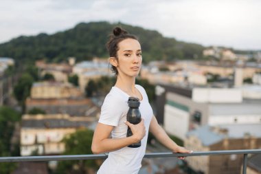 Şehir manzarasına karşı tırabzanların yanında dururken yoga kıyafetleri içinde spor şişesi tutan rahat bir gencin yan görüntüsü. Kendine güvenen bayan sporcu, dış egzersizlerde kullanılan sıvıyı tazeliyor..