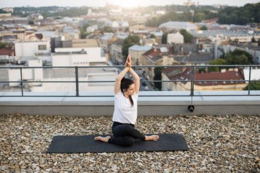 Aktif giysili güzel esmer kadın düz olmayan çatıda elleri bükülmüş bir şekilde Asana egzersizi yapıyordu. Güzel yoga meraklısı, temiz havanın tadını çıkarırken omuzlarındaki esnekliği arttırıyor..