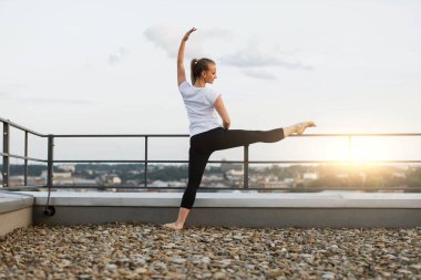 Sarışın bir kadının açık hava eğitiminde bacak kaslarını esnetmek için çatı tırabzanlarını baraj olarak kullanmasının arkası. Aktif giyimde çekici bir insan günbatımının tadını çıkarırken zarafet ve güveni geliştiriyor..