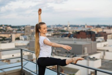 Yoga kıyafetleri içindeki zarif beyaz bayanın binanın çatısının korkuluklarında bacak uzatma hareketi. Profesyonel sporcu şehir manzaralı panoramik terasta bar esneme rutininin keyfini çıkarıyor.