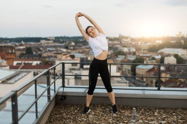 Şehrin arka planında ellerini kaldırarak omurgasını uzatan sakin gencin yakın görüntüsü. Etkileyici spor meraklısı çatıdaki etkin egzersizden sonra omuz kaslarını gevşetiyor..