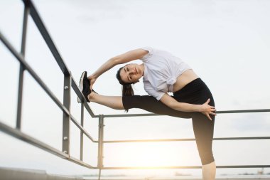 Gün batımında arka planda çatıdaki tırabzanları kullanarak yoga kıyafeti giyen beyaz kadına odaklan. Zarif yetişkin insan kaburga kaslarını güçlendirirken aynı zamanda vücudu hareket ettiriyor..