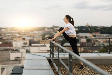 Yaz akşamları şehir manzarasının tadını çıkarırken çatıdaki tırabzanlara bacağını koyan genç bir kadının yan görüntüsü. Atletik esmer bayan panoramik terasta saatlerce süren eğitimden sonra dinleniyor..