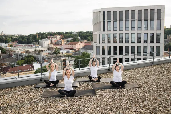 High angle view of young ladies in sportswear taking up sitting meditation pose with raised hands against urban scenery. Beautiful women elevating mind-body connection by practicing yoga outside.