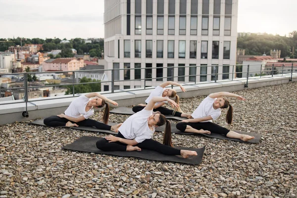 Portrait of charming ladies in activewear exercising spiralled asana on black mats against urban scenery. Positive sportswomen being involved in forward bend with twist during seated yoga practice.