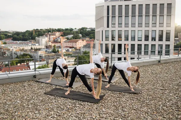 High angle view of women bending aside with one hand touching ankles while reaching left fingertips towards sky. Charming practitioners in yoga outfits exercising on uneven surface of open terrace.