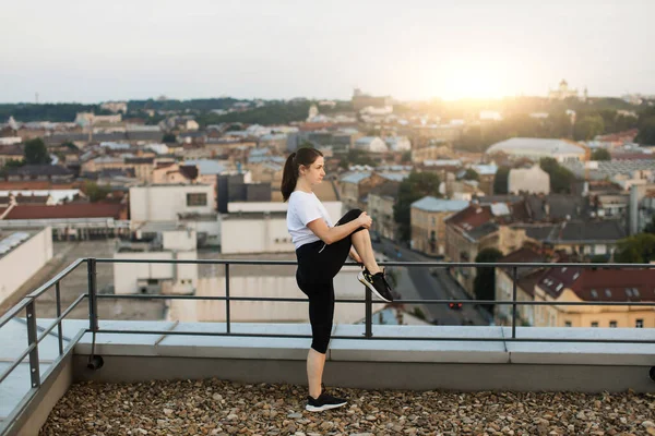 Beautiful brunette female in cozy outfit stretching knees during outdoor warming-up on roof terrace. Young caucasian athlete improving flexibility while starting fitness routine in open air.