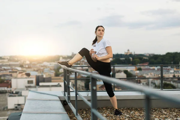 Attractive brunette lady in sports apparel using sports bottle while standing near handrails on gravel roof. Carefree female athlete having rest after training exercises on panoramic terrace.