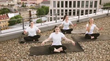 Yoga kıyafetleri içindeki çekici kadınlar panoramik terasta siyah minderlerde otururken yarım nilüfer poziyonuna giriyorlar. Atletik beyaz kadınlar namaste 'yi açık havada klasik oturarak kullanıyorlar..