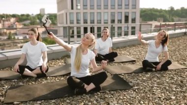 Çekici beyaz kadınlar, yoga minderlerinde bacak bacak bacak üstüne atarak selfie çubuğuyla telefona selam veriyorlar. Atletik yetişkinler panoramik terasta Asya egzersizi yaparken kendi fotoğraflarını çekiyorlar..
