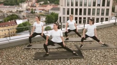 Beyaz tişörtlü ve siyah pantolonlu genç ve zayıf yetişkinler dışarıda vücut duruşu için kaslarını uzatıyor. Güzel kadınlar panoramik terasta dış seans sırasında yoga zihniyetine girerler..