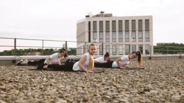 Vücut ölçülerini uzatırken çatıdaki çakıllı araziden göğüslerini soyan spor elbiseli çekici ve fit kadınlara odaklan. Açık hava fitness rutinine devam etmeden önce kısa bir süre yoga pozu veren sporcu kadınlar..