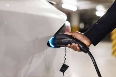 Halka açık otoparkın içindeki modern EV 'nin şarj portuna elektrik prizi takan erkek eli görüntüsü. Resmi olarak giyinmiş bir adam iş yaparken akü sürücüsü arabasına enerji sağlıyor..