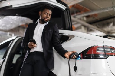 Afrika kökenli Amerikalı erkek, yeraltı otoparkındaki otomobil limanında güç kablosunu tamir ediyor. İyi giyimli profesyonel cep telefonu ile çalışma saatleri ayarlanırken EV şarj oturumu başlıyor.