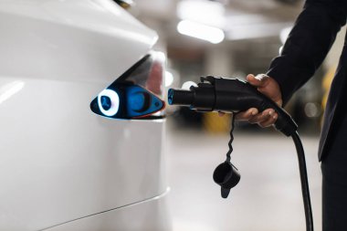 Otoparkın içinde beyaz parlayan elektrikli araba limanına yaklaşan elektrik kablosu olan bir adamın el görüntüsü. Çevre dostu alternatif enerji ve EV şarj altyapısı kavramı.