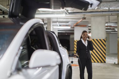 Halka açık garajda saf elektrikli aracı doldururken Afrikalı Amerikalı bir kadının telefonda konuşmasının düşük açılı görüntüsü. Ofis dışındaki şirket meseleleriyle ilgili kararlar veren resmi kıyafetli meşgul bir işçi..