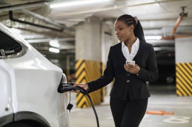 Şarj kablosu olan elektrikli otomobilin yanında duran cep telefonlu çekici bayana odaklan. İş elbisesi içindeki Afrikalı girişimci park yerinde pilli araba hazırlıyor..