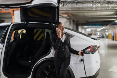 Resmi giysiler içinde gülümseyen bir kadının akıllı bir aletle sohbet ederken e-arabaya enerji sağladığı geniş bir görüş açısı. Neşeli Afrikalı Amerikalı kadın bodrum katındaki park yerinde iletişim yeteneklerini geliştiriyor..