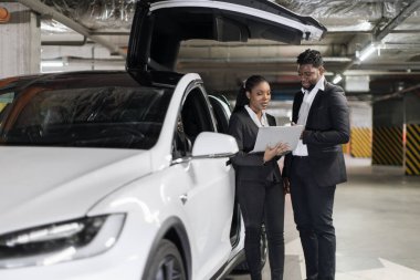Resmi kıyafet giymiş Afrikalı Amerikalı iş ortakları elektronik arabanın yakınındaki dijital ekranda bilgileri izliyorlar. Güzel kadın ve yakışıklı adam iş çıkışı laptopla sunum yapmayı planlıyorlar..