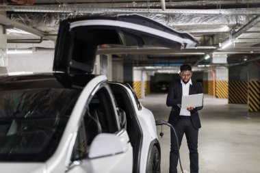 Resmi kıyafetli Afrikalı iş adamının seçici odak noktası EV otoparkında taşınabilir dizüstü bilgisayar kullanması. E- araba yakınlarında ofis duvarları dışında profesyonel faaliyet yürüten nitelikli bir çalışan..