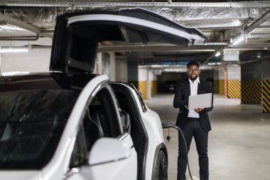 Resmi kıyafetli Afrikalı iş adamının seçici odak noktası EV otoparkında taşınabilir dizüstü bilgisayar kullanması. E- araba yakınlarında ofis duvarları dışında profesyonel faaliyet yürüten nitelikli bir çalışan..