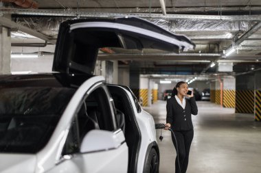 Resmi olarak neşeli genç bir kadın EV otoparkında şarj kablosu tutarken akıllı aletlerle sohbet ediyor. Gülümseyen Afrikalı yetişkin, gelecekteki iş toplantısının ana unsurlarını meslektaşlarına özetliyor.