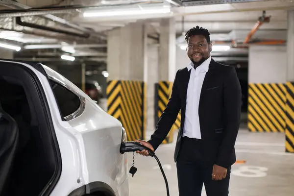 Portrait of middle-aged man in formal outfit holding charging cord connected to electric car in basement parking. African american office worker keeping auto plugged in during work in business center.