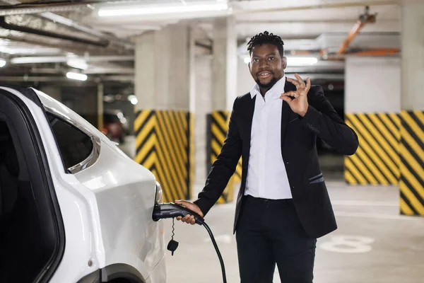Portrait of middle-aged man in formal outfit holding charging cord connected to electric car in basement parking, showing sign ok