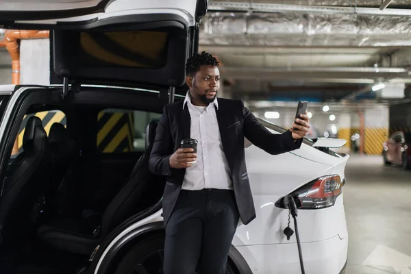 African office worker with beverage taking selfie on cell phone while taking relaxed pose near EV on recharge in garage. Thoughtful employee testing mobile application in public basement parking.