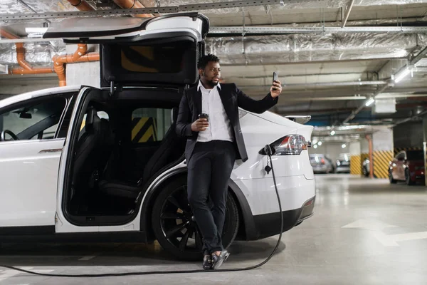 Full length view of attractive man in business suit taking photo on smartphone while having coffee near e-car on fill-up. African american employee catching moment of relaxition in parking area.