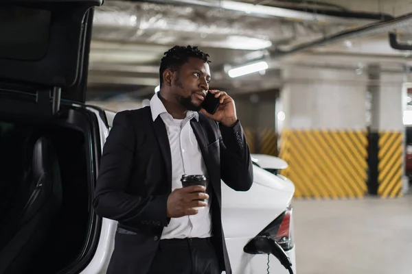 African american person with beverage receiving phone call while waiting for e-car being recharged in office building. Relaxed middle-aged driver in formal suit listening to business report in parking