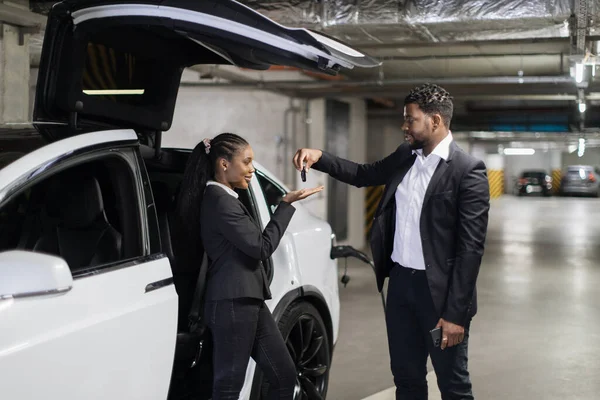 African american man in formal attire giving EV keys to business lady at pick-up location in underground parking. Female customer renting electric auto from charge point after signing agreement.