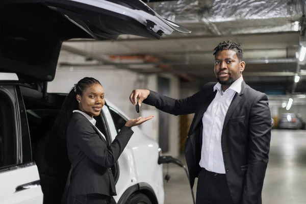 Portrait of african man and woman with smartphones posing near EV in charging session during rental procedure. Smiling car company manager handing over auto keys to new client in basement garage.