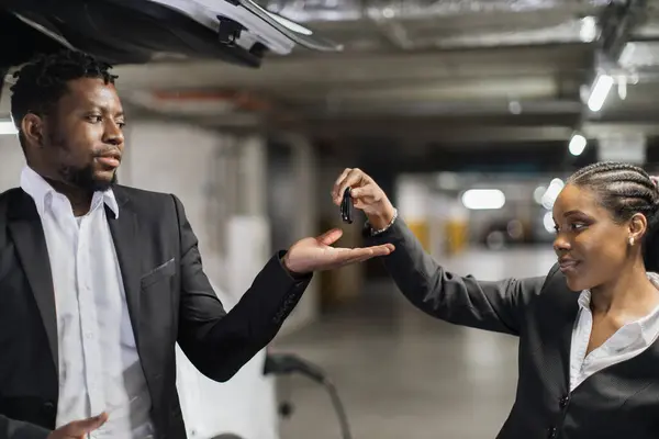 Close up view of african american man and woman being involved in auto keys transfer procedure on car park background. Handsome adult picking up brand new electric vehicle from EV charge point.