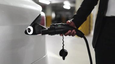 Halka açık otoparkın içindeki modern EV 'nin şarj portuna elektrik prizi takan erkek eli görüntüsü. Resmi olarak giyinmiş bir adam iş yaparken akü sürücüsü arabasına enerji sağlıyor..