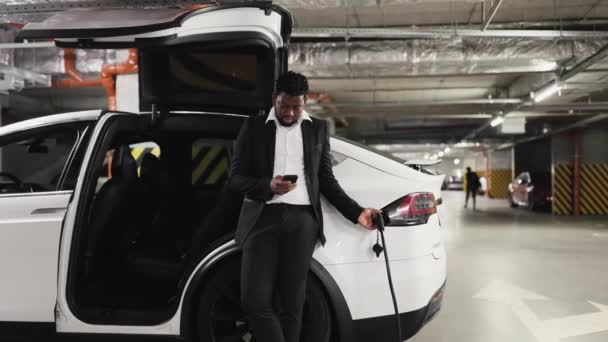 公共駐車場のバッテリー駆動車に適切なコネクターを接続する自信のあるビジネスマン 電気自動車を充電するためのモバイルアプリを使用して正式な衣装のアフリカ系アメリカ人 — ストック動画
