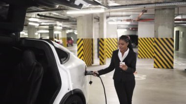 Resmi giyinmiş Afrikalı Amerikalı bir kadın otomotiv portuna elektrik kablosu takarken mobil ekranı tarıyor. EV garajda tam şarja ulaşırken, odaklanmış çalışan mesajları kontrol ediyor.